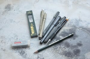 A collection of pens, pencils, and erasers for artistic and design purposes on a textured surface.